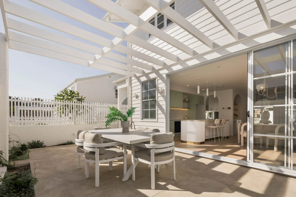 White outdoor dining area under a slatted pergola beside a modern kitchen with glass doors, table and cushioned chairs, and potted plants.