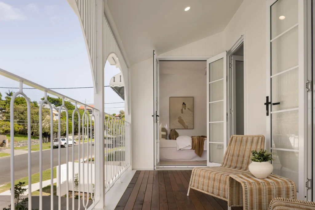 White porch with open French doors leading to a bright bedroom; wicker chairs on a wooden deck with a potted plant in the foreground and a fenced street view beyond.