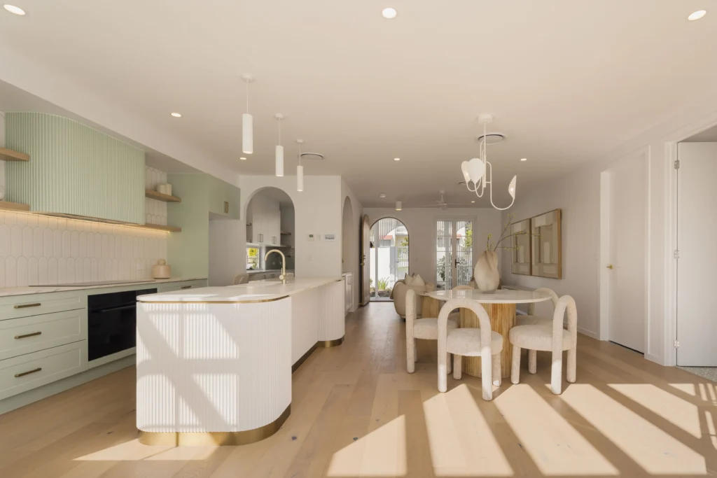 Open-plan kitchen and dining area with pastel green cabinetry, a curved white ribbed island, and a round dining table with beige chairs in natural light.