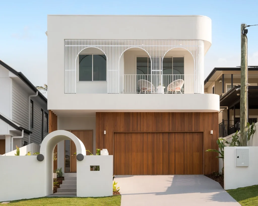 Modern white multi‑story house with arched balcony railings, large windows, and a wood-paneled lower façade; circular sculpture in front.