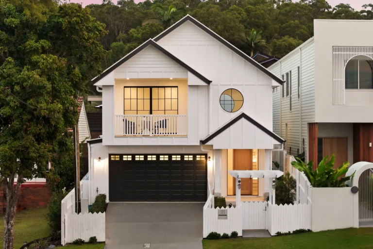 White two-story house with black garage doors, triangular gables, a round window, balcony, and white picket fence along a gated driveway.