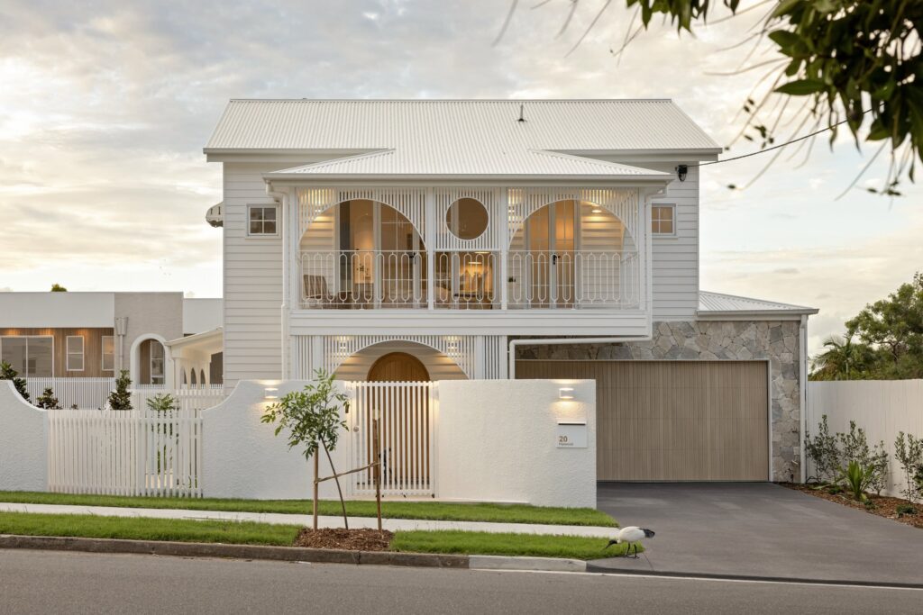 White two-story house with a decorative lattice balcony, circular porch openings, and a gated front yard on a quiet street.